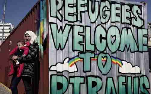 Refugee mother and child stand next to a large sign welcoming refugees ferried from Lesbos to the Greek port of Piraeus